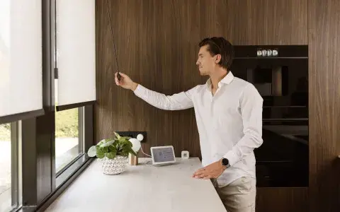 A man raising a MotionBlinds roller blind with a remote in a timber-panelled kitchen, MotionBlinds by Coulisse supplied by Custom Blinds Shutters & Awnings