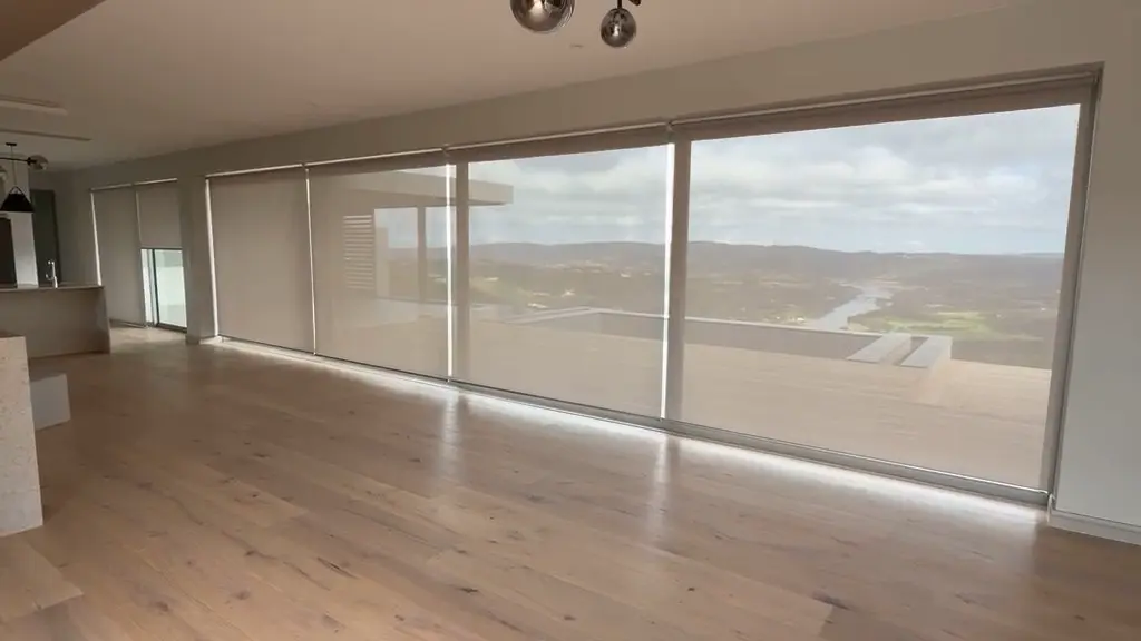 Motorised sheer roller blinds lowered across a full-width glass wall with a river valley view, installed by Custom Blinds Shutters & Awnings