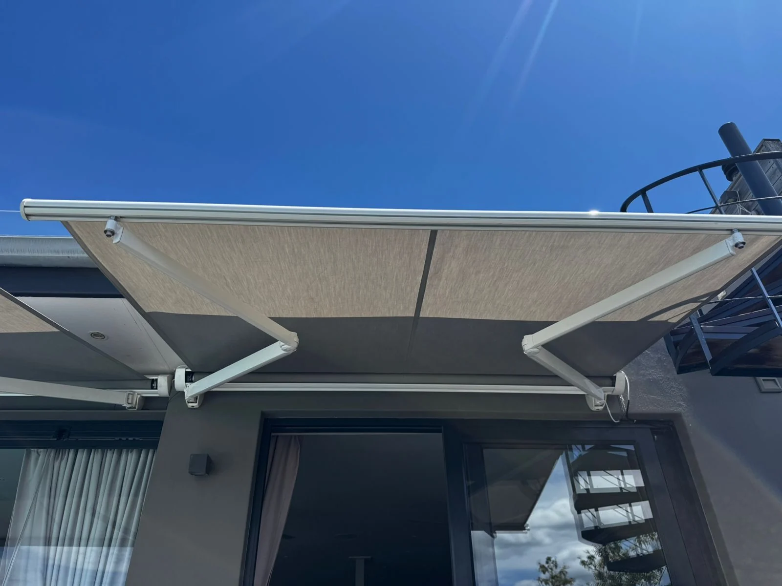 A retractable folding-arm awning by Custom Blinds Shutters & Awnings with beige fabric and white arms, extended over sliding doors against a blue sky, Plettenberg Bay, Garden Route, South Africa