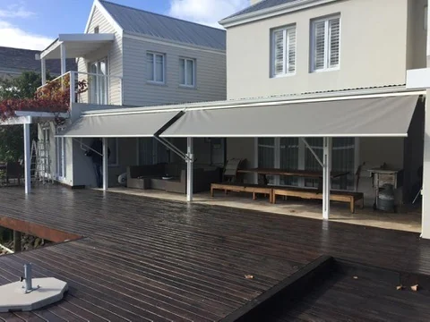 Two grey folding-arm awnings by Custom Blinds Shutters & Awnings extended side by side over a dark timber deck in front of a white weatherboard house, Thesen Island, Knysna, Garden Route, South Africa