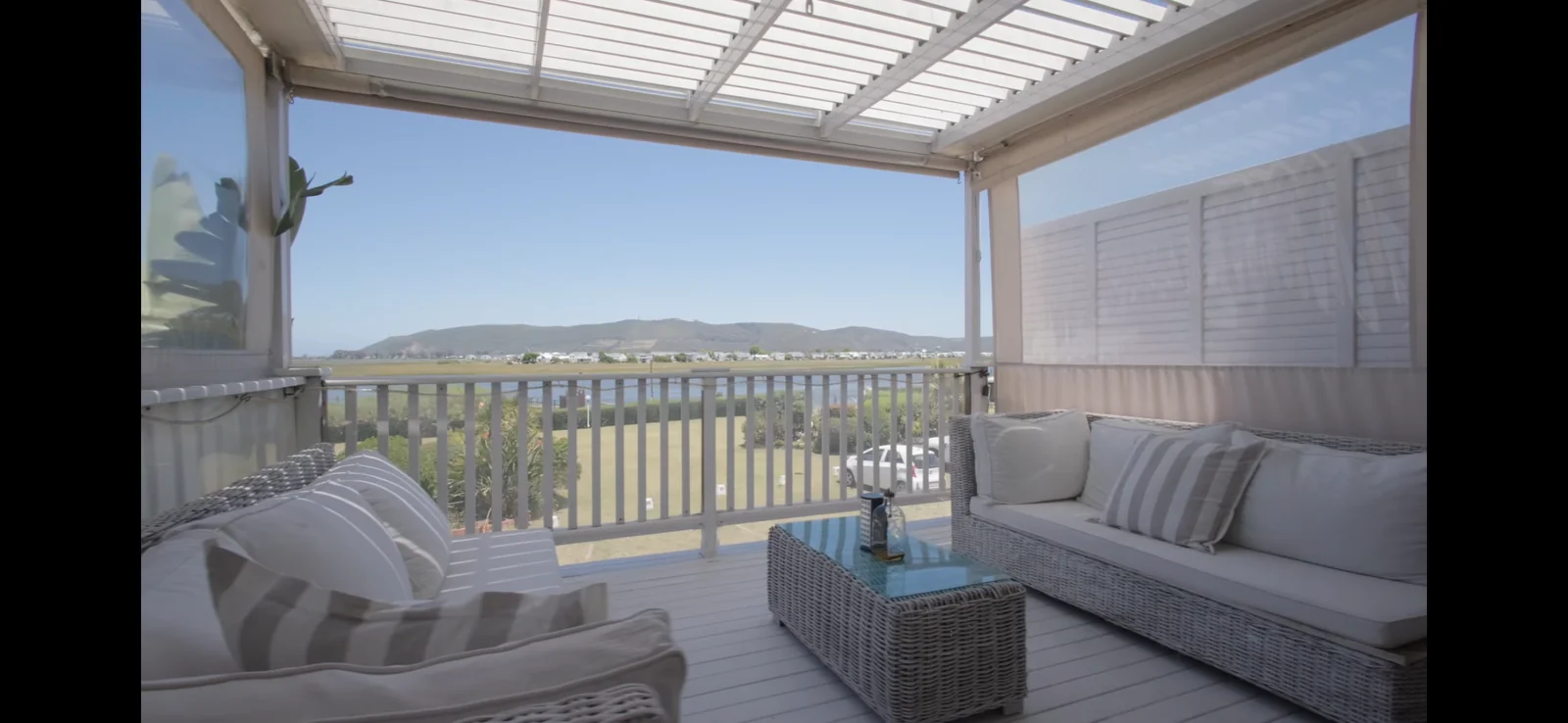 A white louvred awning and outdoor blinds by Custom Blinds Shutters & Awnings over a raised deck with wicker seating, blades open above an estuary view, Costa Sarda, Knysna, Garden Route, South Africa