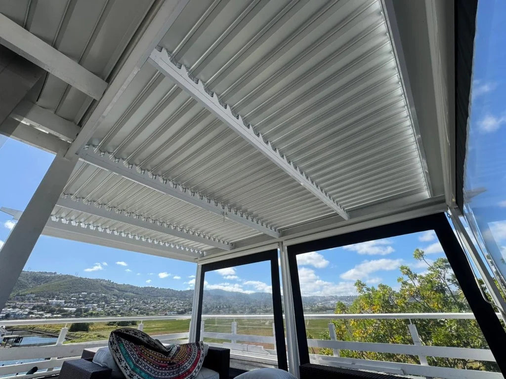 A white louvre awning by Custom Blinds Shutters & Awnings over a deck with outdoor blinds at its edges, blades open above a lagoon and hillside town, Thesen Island, Knysna, Garden Route, South Africa