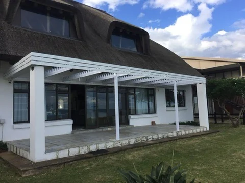 A white louvre awning by Custom Blinds Shutters & Awnings on a paved patio in front of a thatched house, blades open, Herolds Bay, Garden Route, South Africa
