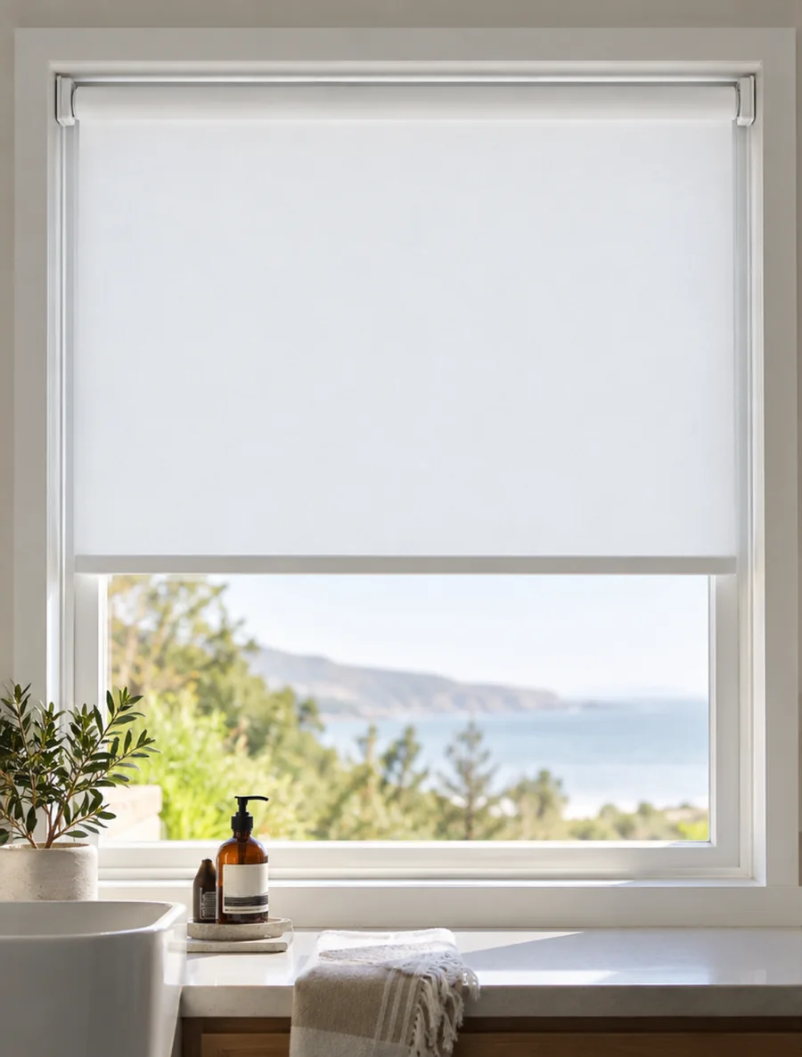 White no-drill roller blind half lowered in a bathroom window above a basin, sea and hills beyond, from Custom Blinds Shutters & Awnings