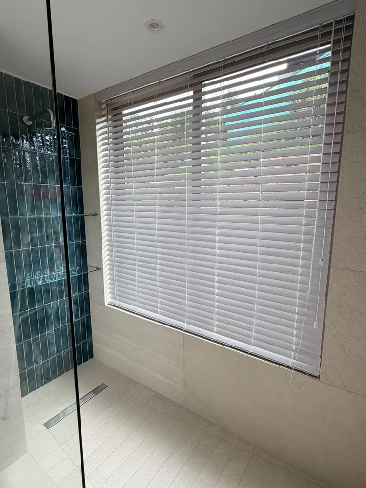White wood-look venetian blinds in a tiled bathroom beside a walk-in shower, fitted by Custom Blinds Shutters & Awnings
