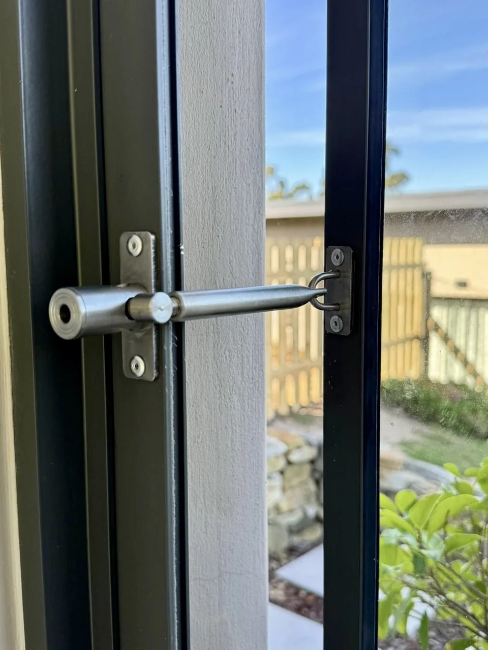 A LockLatch safety latch fitted to an aluminium door by Custom Blinds Shutters & Awnings, the stainless latch arm engaged in its keeper with the garden beyond