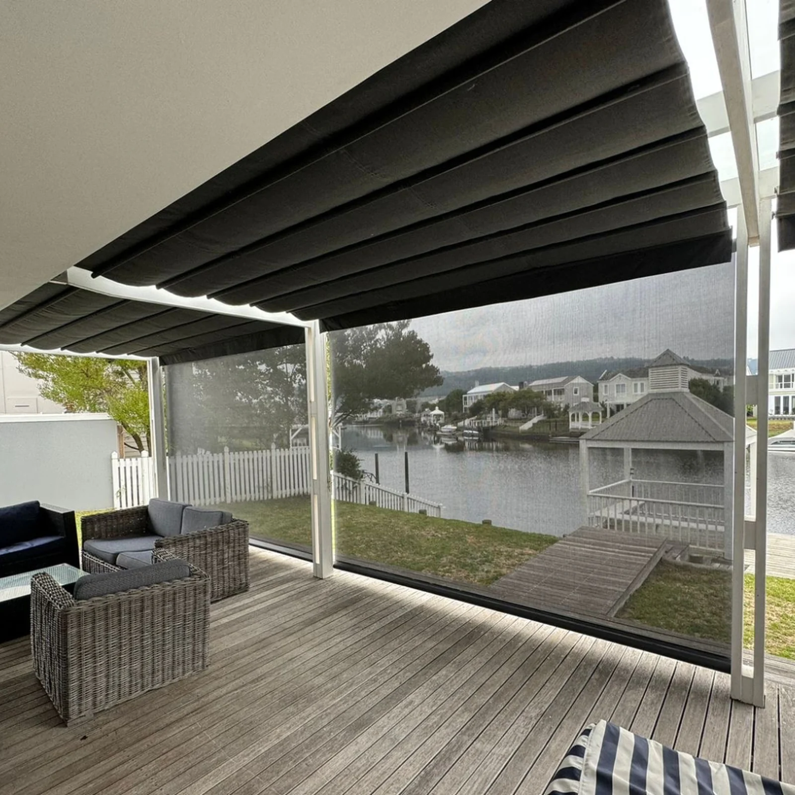 Screen mesh outdoor blinds lowered along a covered timber deck, the lagoon, jetty and houses on the far bank visible straight through the weave, by Custom Blinds Shutters & Awnings, Garden Route, South Africa