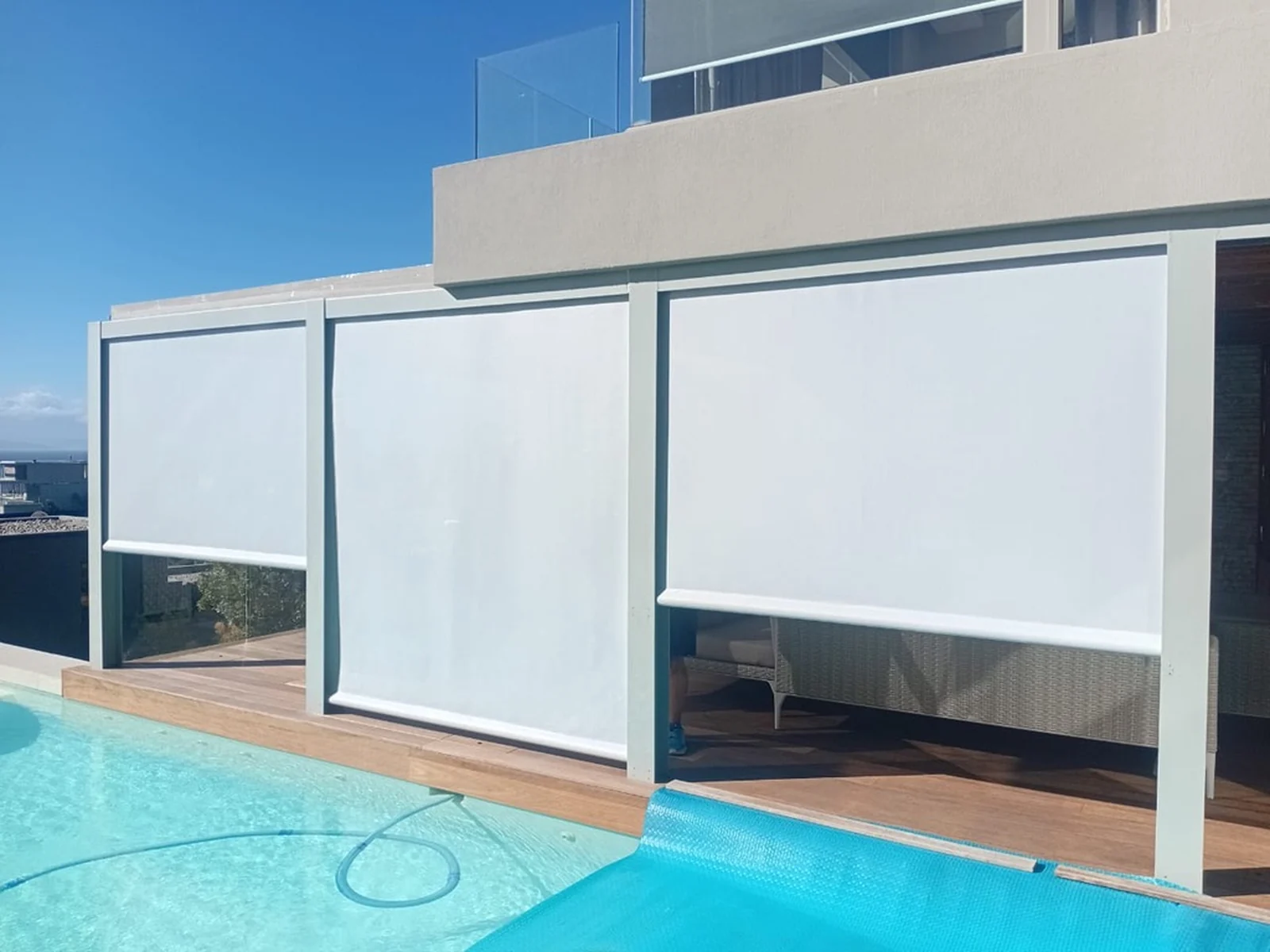 Three white screen blinds lowered in side tracks along a poolside deck, running between white posts under an upper balcony by Custom Blinds Shutters & Awnings, Garden Route, South Africa