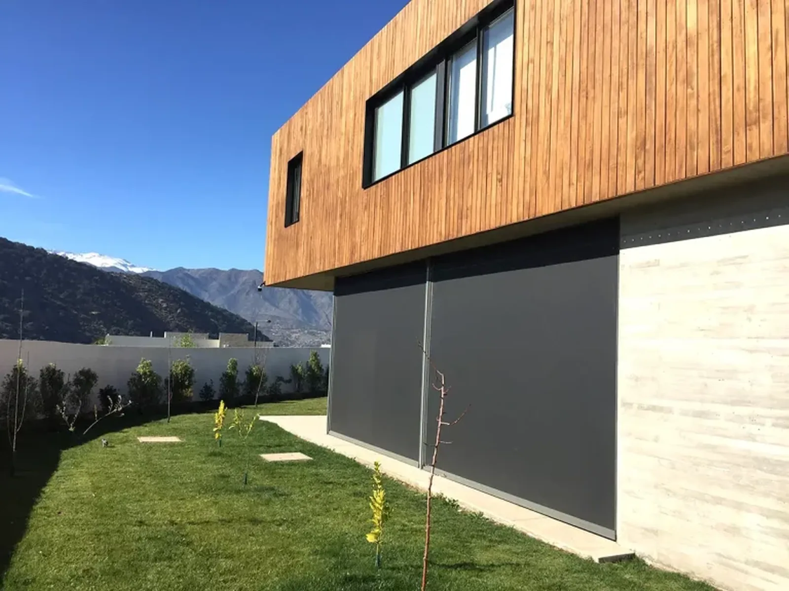 Track guided outdoor blinds lowered across two bays of a timber clad home, aluminium side tracks visible between the bays, mountains beyond, by Custom Blinds Shutters and Awnings