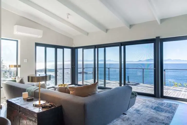 Black-framed sliding security screens across a lounge opening onto a deck with a sea view, by Custom Blinds Shutters & Awnings