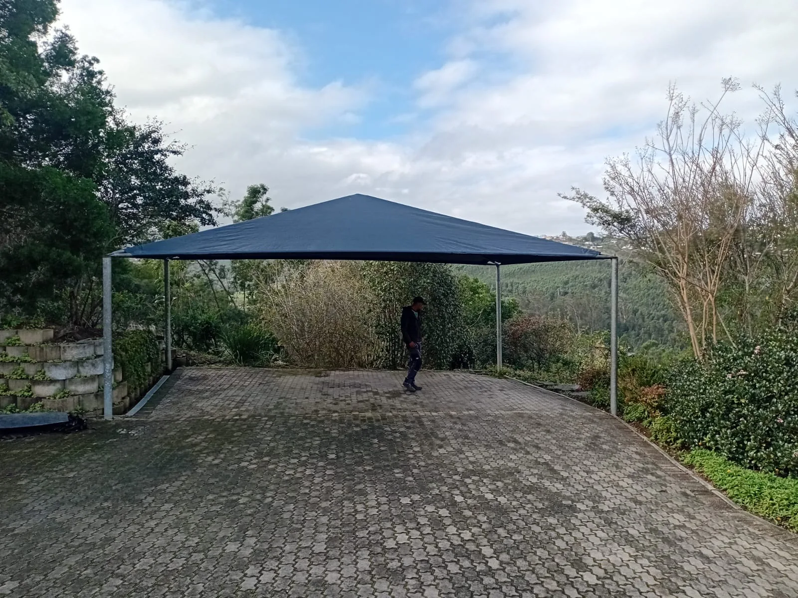 A charcoal four post shadeport by Custom Blinds Shutters & Awnings, its shade cloth canopy spanning a wide paved driveway with forested hills and the Garden Route valley behind