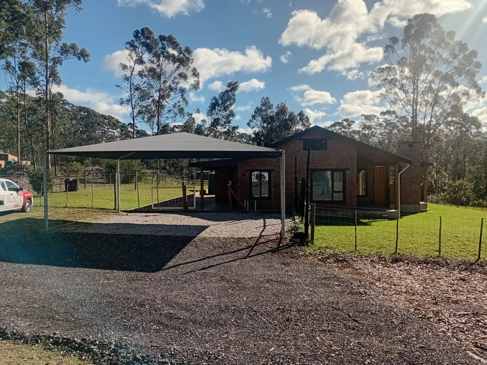 A grey four post shadeport installed by Custom Blinds Shutters & Awnings on a gravel driveway beside a face brick Garden Route home, the Custom Blinds Shutters & Awnings vehicle parked alongside