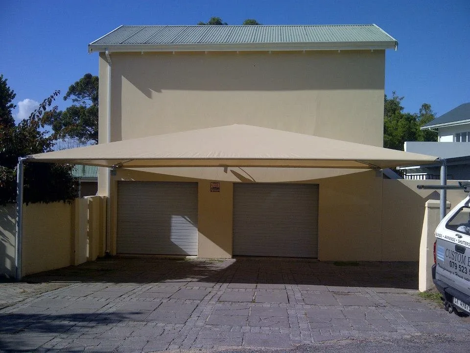A cream shadeport by Custom Blinds Shutters & Awnings extending from a double garage over a brick paved driveway, the Custom Blinds Shutters & Awnings vehicle at the kerb