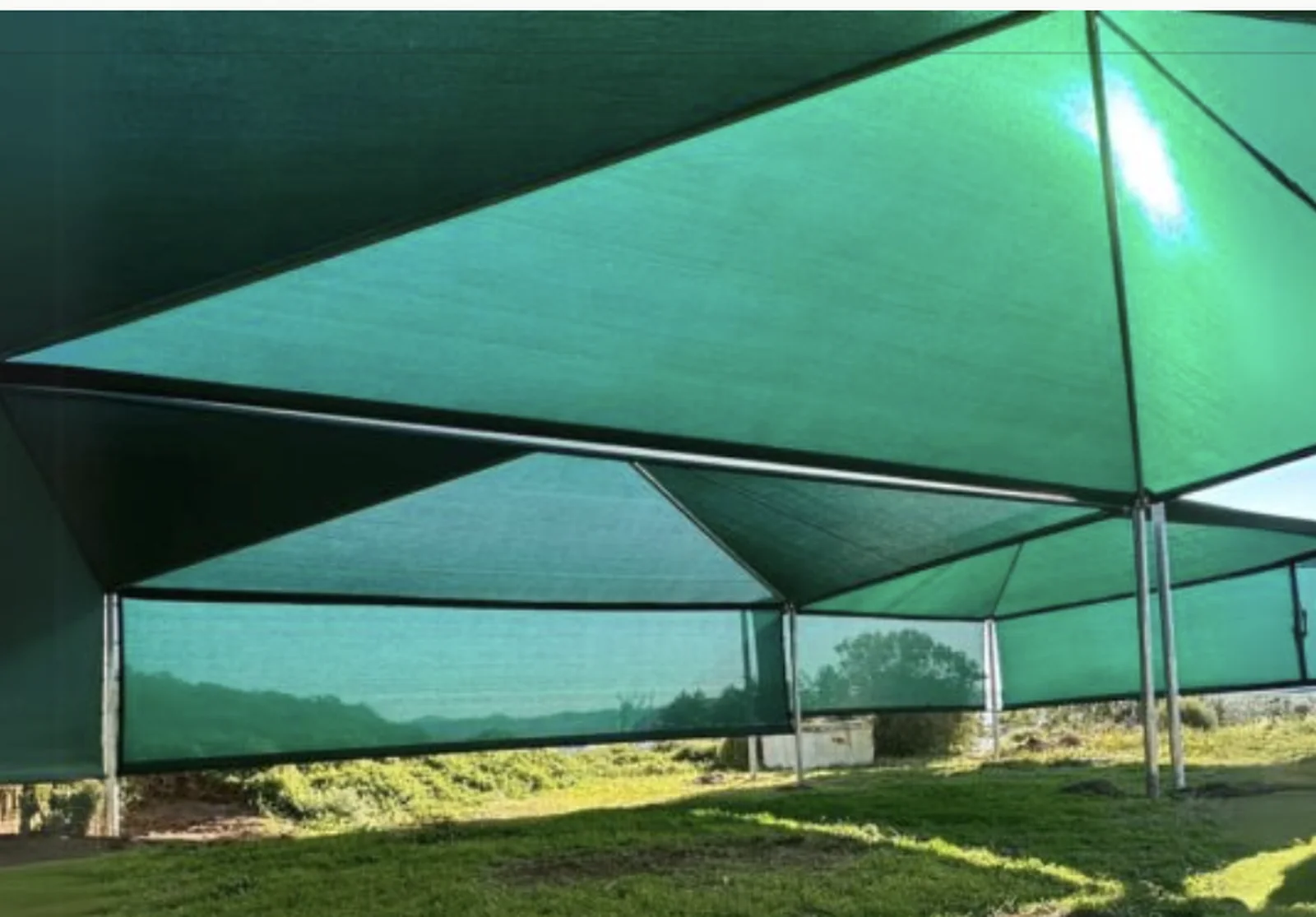 A large green shade cloth shadeport by Custom Blinds Shutters & Awnings seen from underneath, the tensioned canopy panels running away over a grassed area with side drops and a valley beyond