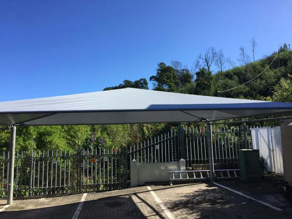 A pale grey shadeport by Custom Blinds Shutters & Awnings over marked parking bays at a Garden Route complex, palisade fencing and indigenous bush behind