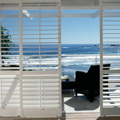 White PVC shutters by Custom Blinds Shutters & Awnings folded back across a coastal living room's glass doors, louvres open to the sea
