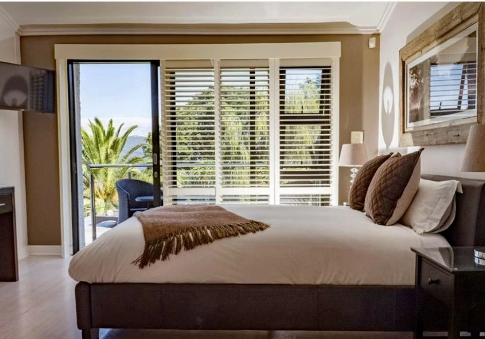 PVC indoor shutters in a guest bedroom at Cambalala Guesthouse, Knysna, lagoon views through the open balcony door, by Custom Blinds Shutters & Awnings