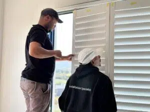 Custom Blinds South Africa Two people fit white window shutters in a sunny home, making the space more private. One wears a shirt saying “Installation Specialist”. Garden Route & Online
