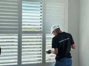 Custom Blinds South Africa An installer in a white cap adjusts shutters in a sunlit home, letting in light whilst keeping the room cool and comfortable. George Garden Route & Online