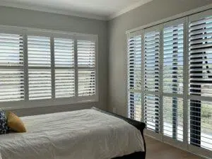 Custom Blinds South Africa Sunny bedroom with large windows and white shutters, light floors, and fresh linen. Shutters let in light whilst giving privacy. George Garden Route & Online