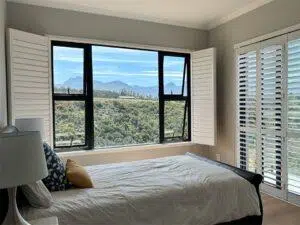Custom Blinds South Africa Sunny bedroom with white shutters and a big window, framing peaceful hills. Bright bed linen bring life and privacy to your space. George Garden Route & Online