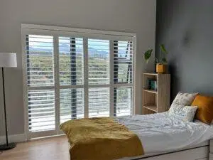 Custom Blinds South Africa Bright bedroom with a single bed, mustard bedding, wooden shelf, and white shutters letting in mountain views—fresh and inviting. George Garden Route & Online
