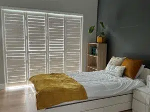 Custom Blinds South Africa A cosy bedroom with soft white linen, mustard touches, and large shutters brightening the room—perfect for relaxed South African living. George Garden Route & Online