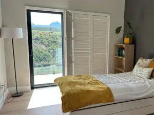 Custom Blinds South Africa Sunny bedroom with a cosy bed, bookshelves, and plants. Glass doors reveal mountain views, filling the room with fresh light and calm.George Garden Route & Online