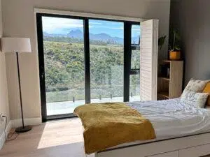 Custom Blinds South Africa Sunny bedroom with a bed, yellow throw, bookcase and lamp. Glass doors reveal a balcony and views of green hills and distant peaks. George Garden Route & Online