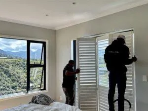 Custom Blinds South Africa Two custom blinds installers fit white shutters in a cosy bedroom, letting in views of rolling hills whilst adding privacy and a fresh, tidy look. Garden Route & Online