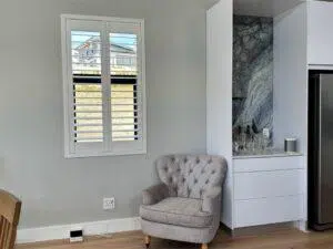 Custom Blinds South Africa A comfy grey chair by a neat cupboard and window with white shutters, brightening up the lounge with light floors and soft grey walls. George Garden Route & Online