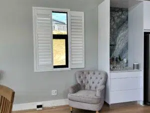Custom Blinds South Africa A cosy grey armchair brightens this lounge, whilst white shutters let in light. A stylish cabinet neatly displays glassware. after custom blinds shutters installed in George Garden Route & Online