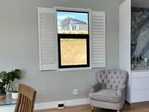 Custom Blinds South Africa A comfy grey armchair by white shutters lets in garden views; perfect for relaxing, with a homely touch from plants and glassware. Garden Route & Online