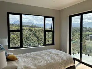Custom Blinds South Africa Sunlight streams into this airy bedroom, where large windows frame green hills—perfect for a tranquil, refreshed space. before custom blinds shutters Garden Route & Online