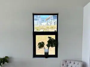 Custom Blinds South Africa A black-framed window brightens a light grey wall; greenery outside and a cosy spot inside create an inviting, stylish home. before custom blinds shutters Garden Route & Online