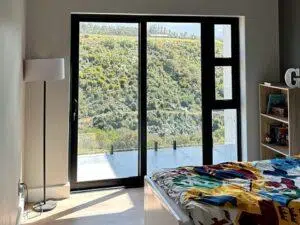 Custom Blinds South Africa Sunny bedroom with glass doors to the balcony, stunning views of green hills, vibrant bedding, and neat built-in shelving. before image for our internal shutters Garden Route & Online
