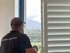 Custom Blinds South Africa An expert in a black Installation Specialist shirt admires the scenic view through sleek white blinds, brightening up the George home. Garden Route & Online