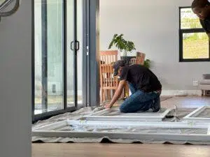Custom Blinds South Africa A Custom blinds installer fits a new white window frame, brightening a stylish home. Large glass doors connect the space to light and fresh air. Garden Route & Online