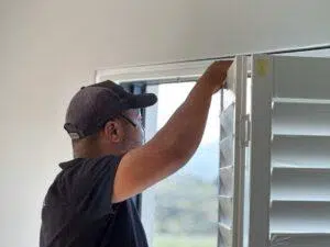 Custom Blinds South Africa Wearing a black cap, a person fits white shutters to brighten the room and control sunlight for a more comfortable home. Garden Route & Online