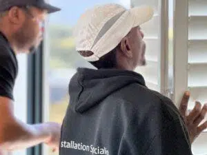 Custom Blinds South Africa Two men, one in a white cap and black hoodie, adjust white shutters indoors for better light and privacy in the home. Garden Route & Online