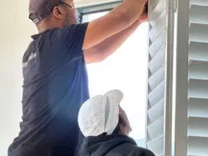 Custom Blinds South Africa Two Custom Blinds installers fit white shutters indoors, making the space brighter and more comfortable. Natural light streams through the window in George . Garden Route & Online