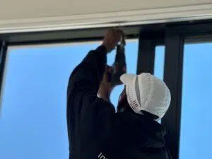 Custom Blinds South Africa a Custom Blinds installer in a white cap and black hoodie drills into a window frame, letting fresh light stream into the room under bright blue skies. Garden Route & Online