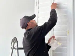 Custom Blinds South Africa A Custom Blinds installer on a stepladder paints white window shutters, brightening the home and protecting from harsh sun with a fresh new look. George Garden Route & Online