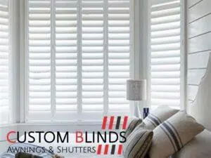 Custom Blinds Shutters & Awnings South Africa Sunny room with white shutters, striped cushion, and cosy lamp. Blinds from Custom add both style and privacy to your space. Garden Route & Online