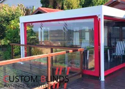 Custom Blinds Shutters & Awnings South Africa A stylish deck with glass balustrades and PVC roller blinds creates a comfy Knysna patio. Custom Blinds Awnings & Shutters logo in the corner. Garden Route & Online