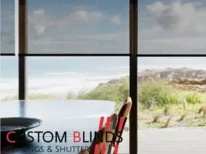 Custom Blinds Shutters & Awnings South Africa Spacious dining room with easy-care blinds frames seaside views, perfect for effortless upkeep in holiday homes. Brand logo in corner. Garden Route & Online