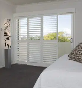 Custom Blinds South Africa Sunny bedroom in George home with a comfy bed and indoor plantation shutters that let in light, soften glare, and frame leafy outdoor views. Garden Route & Online