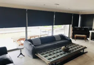 Custom Blinds Shutters & Awnings South Africa A cosy lounge with blockout grey roller blinds controlling the sun, comfy sofa, rustic table, piano and outside seating for laid-back living. Garden Route & Online