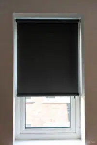 Custom Blinds Shutters & Awnings South Africa A white-framed window with a black roller blind and no drill adaptions blocks glare, whilst letting in light and blurred views of the outside brick building. Garden Route & Online