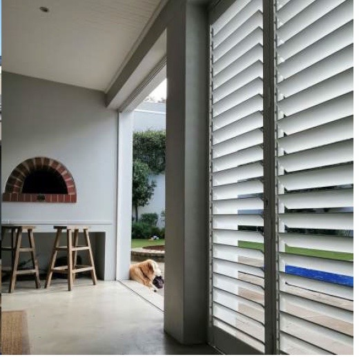 Custom Blinds Shutters & Awnings South Africa Indoor space with white plantation shutters, brick pizza oven, and stools; doors open to garden where shutters let light in and dog relaxes. Garden Route & Online Custom Blinds Shutters & Awnings South Africa Indoor space with white plantation shutters, brick pizza oven, and stools; doors open to garden where shutters let light in and dog relaxes. Garden Route & Online